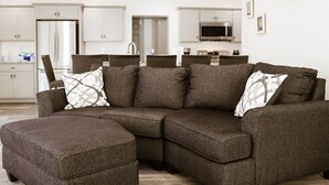 Een moderne woonkamer met een hoekbank, een poef en een keuken op de achtergrond.