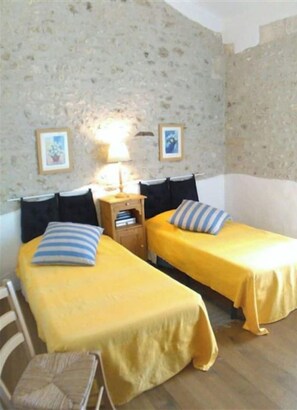 Two single beds with yellow bedspreads and blue-striped pillows, situated against a stone wall with framed pictures above.
