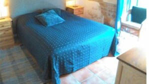 Een slaapkamer met een stenen muur, een bed met een blauw beddegoed, een houten hoofdeinde, een nachtkastje met een lamp en een raam met gordijnen.