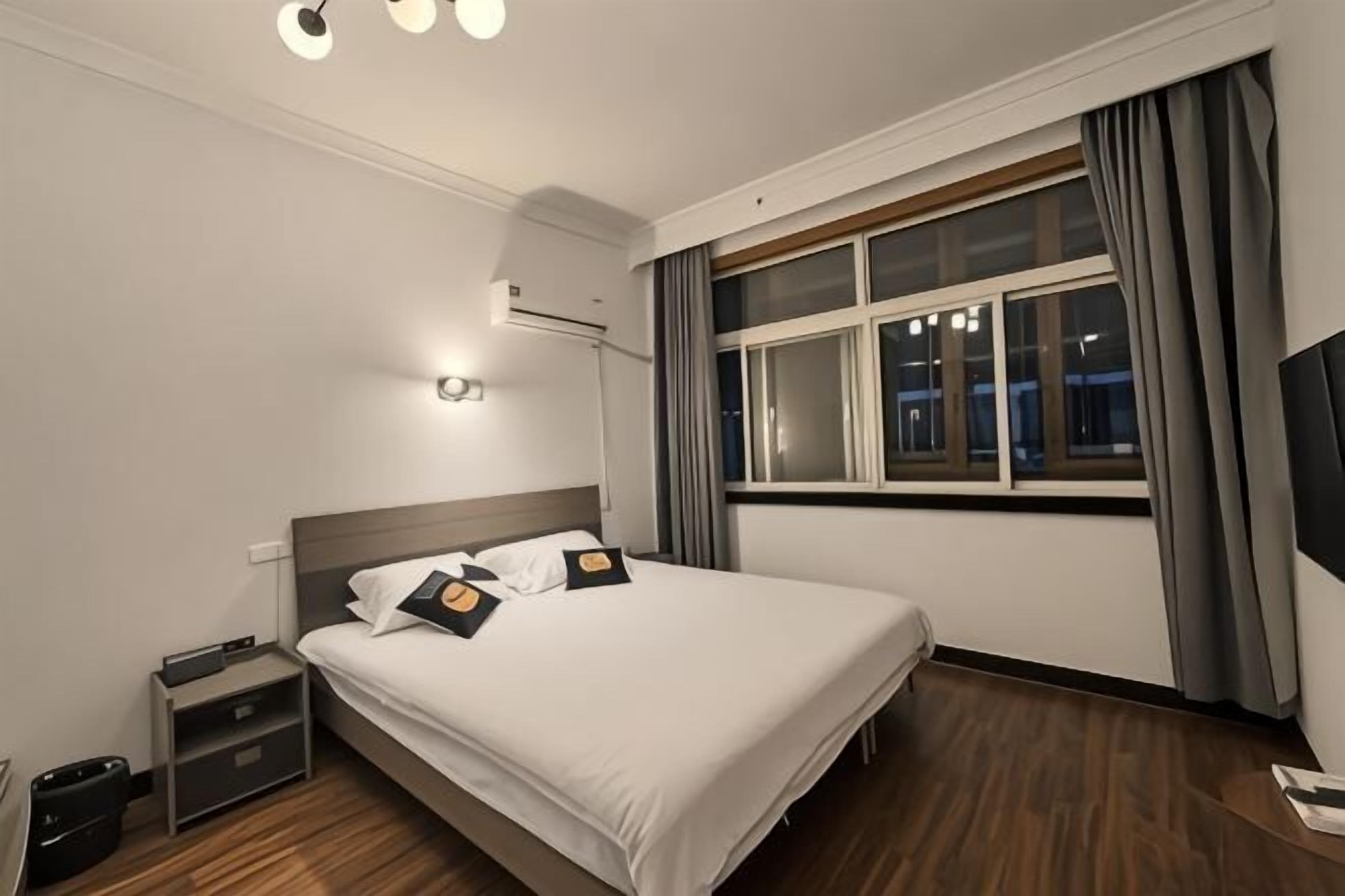 A hotel room with a bed, bedside table, lamp, window with curtains, and a wall-mounted TV.