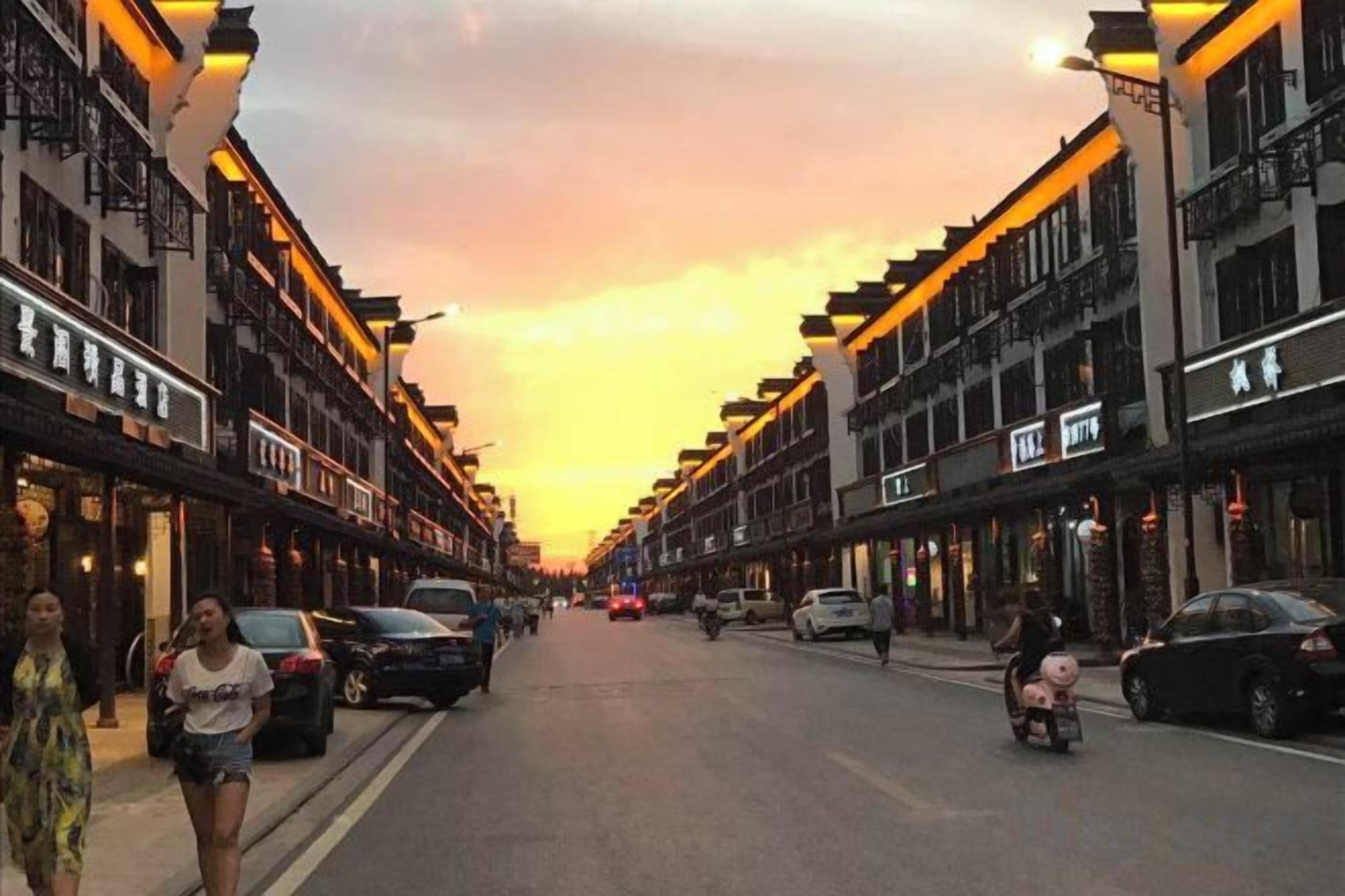A street lined with buildings, cars parked along the sides, and a sunset in the background.