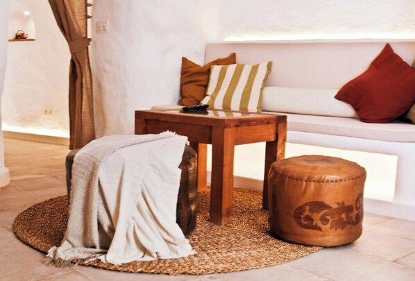 Ein gemütliches Zimmer mit einer weißen Couch, einem Holznachttisch, einem gewebten Teppich und einem Lederhocker.