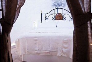 Uma cama com cabeceira de madeira, roupa de cama branca e vista para uma árvore através de uma janela.
