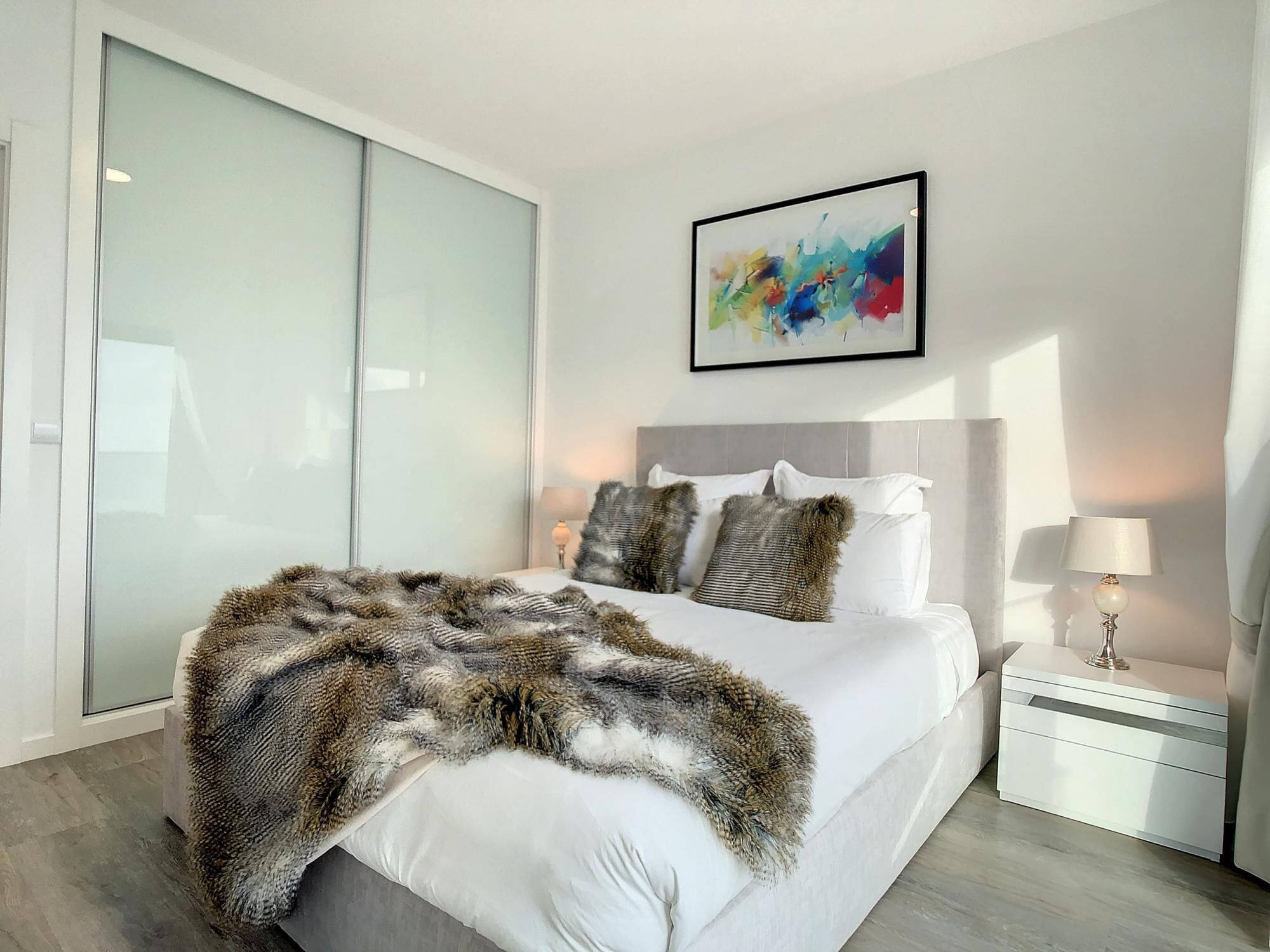 Une chambre moderne avec un grand lit, une armoire en verre, un tableau abstrait aux couleurs vives et une table de chevet avec une lampe.