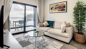 Uma sala de estar moderna com um sofá, uma mesa de centro de vidro e vista para o mar através de grandes janelas.