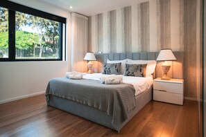 Un dormitorio moderno con una cama grande, dos mesitas de noche con lámparas, vistas a zonas verdes a través de una ventana y una pared a rayas.
