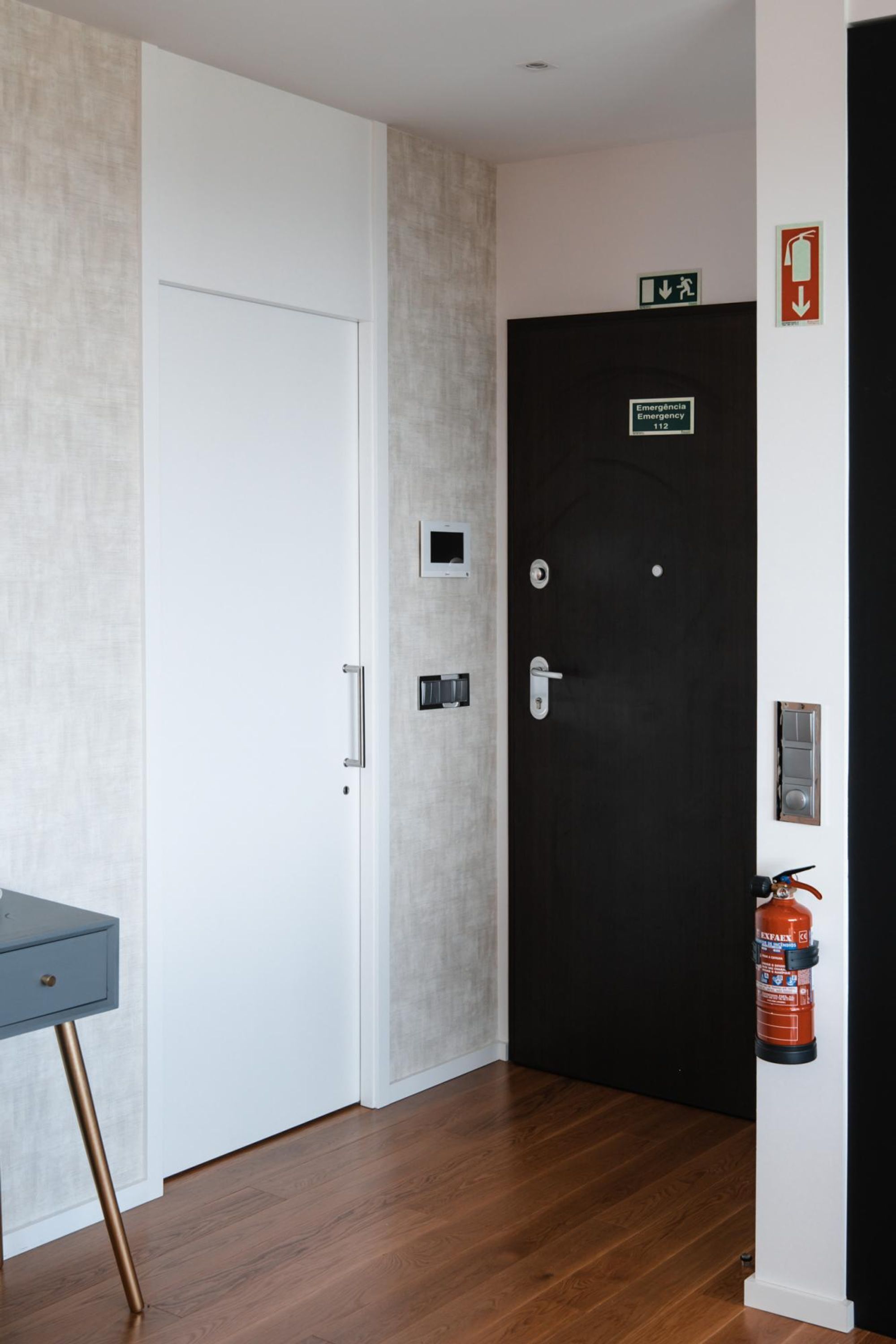 Un pasillo con una puerta blanca, una puerta negra con letrero de salida de emergencia, un extintor y una pequeña mesa con un cajón.