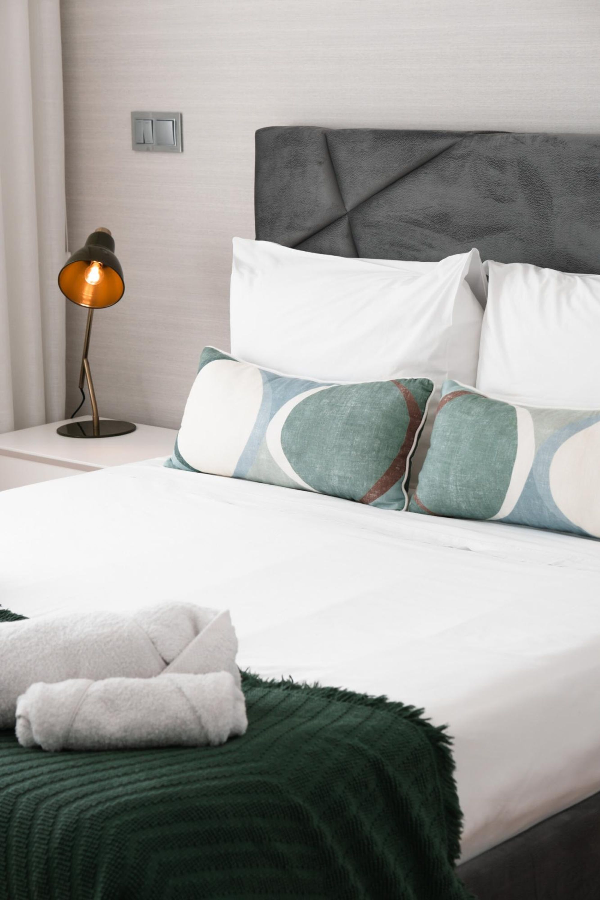 Una cama bien hecha con ropa blanca y almohadas decorativas, una manta verde y una lámpara de noche.