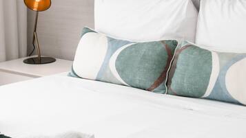Una cama bien hecha con ropa blanca y almohadas decorativas, una manta verde y una lámpara de noche.