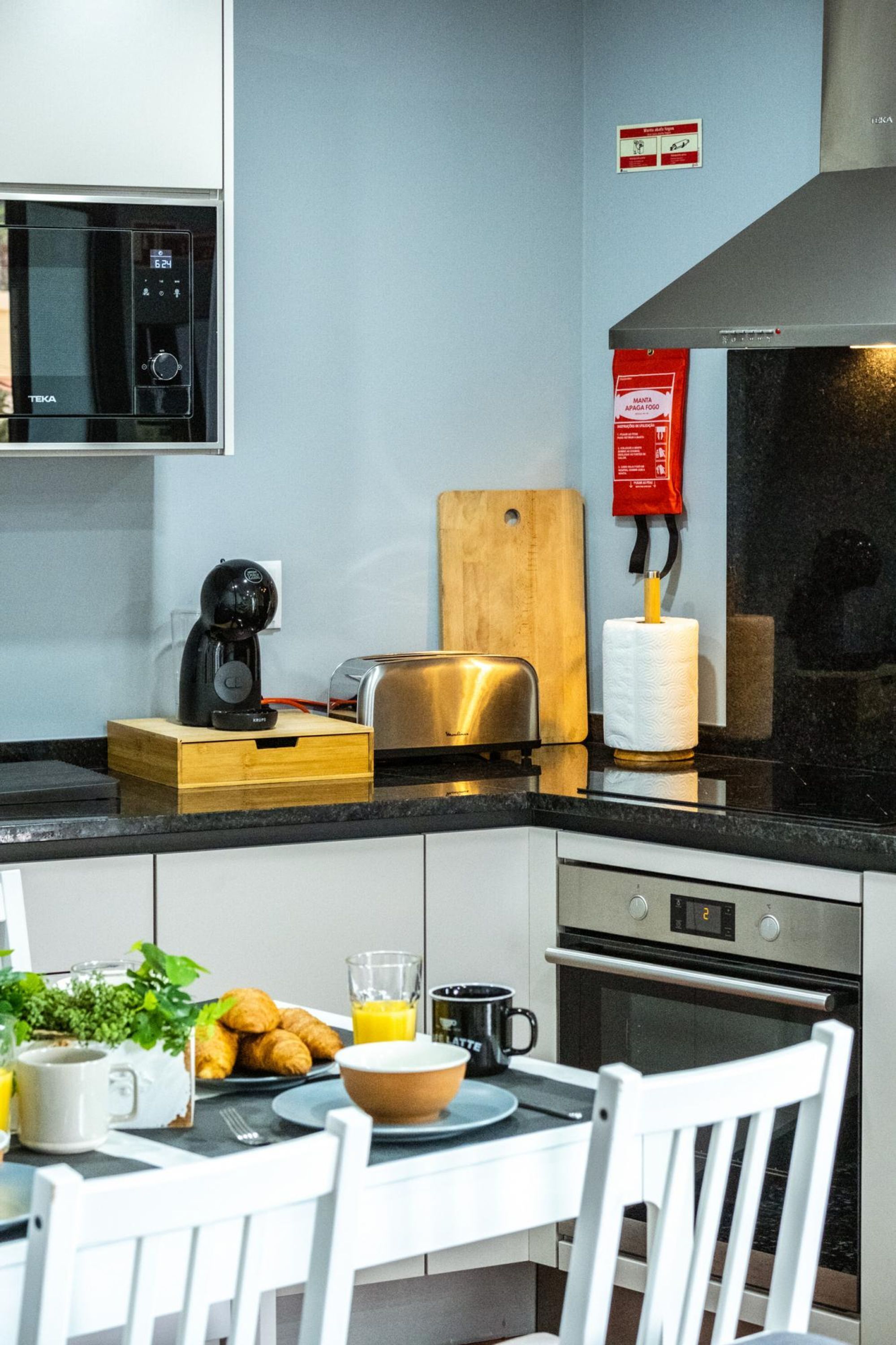 A modern kitchen with a microwave, toaster, and wooden cutting board. There is a coffee maker, a roll of paper towels, and a bowl of food on the counter. A chair and a table are also visible.