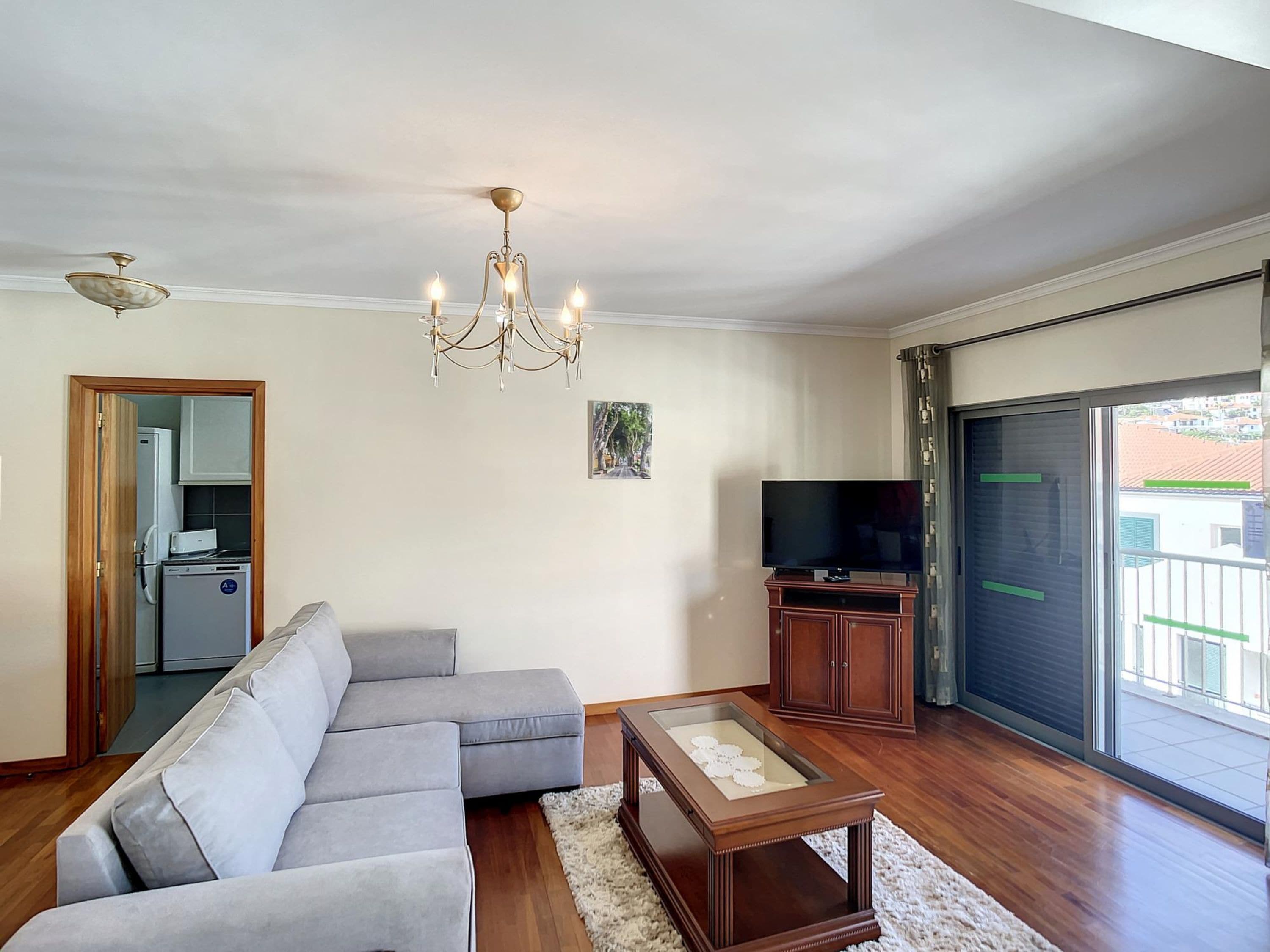 Una sala de estar con un sofá seccional gris, una mesa de centro de madera, un televisor sobre un mueble de madera, una araña y un ventanal corredizo al exterior.