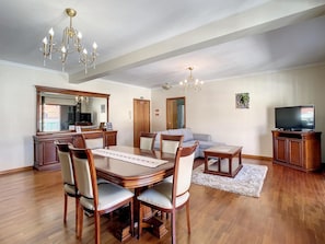 A spacious living room with a wooden dining table, chairs, a sofa, and a TV. There are chandeliers and a large mirror above the table.