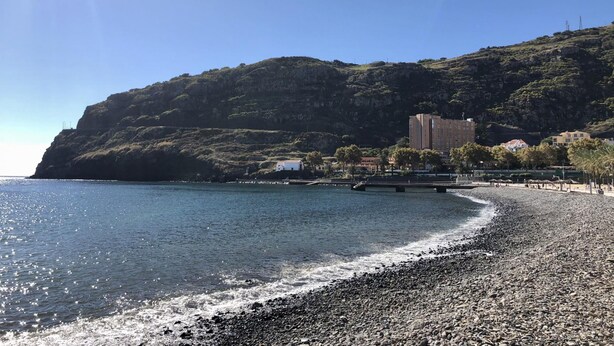 Una playa costera con un litoral rocoso, cielo azul despejado y un edificio en una colina al fondo.