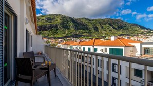 Un balcón con sillas de mimbre y una mesa, con vista a un pueblo de edificios con tejados rojos y colinas verdes.