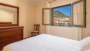 Una habitación de hotel con una cama, un aparador de madera, una silla y una ventana con vistas a un pueblo y montañas.