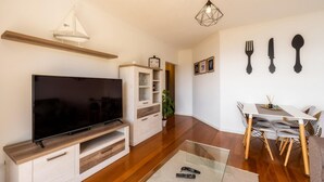 A living room with a flat-screen TV, wooden flooring, a dining area with a table and chairs, and wall-mounted cutlery. - Sea View by Atlantic Holiday (Santa Cruz)