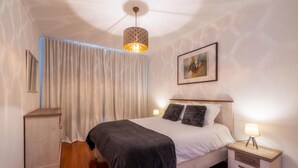 A bedroom with a bed, bedside tables, a nightstand, a hanging light fixture, a window with curtains, and a wall-mounted picture. - Sea View by Atlantic Holiday (Santa Cruz)
