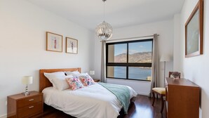 A bedroom with a wooden bed, a nightstand, a desk, a chair, a large window with a view of the sea, and a chandelier. - Centromar Residence Ocean View by Atlantic Holiday (Funchal)