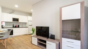 Une cuisine moderne avec des meubles blancs, un four à micro-ondes et un réfrigérateur. On y trouve un coin repas avec une table et des chaises. Un téléviseur est posé sur un meuble en bois.