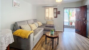 A living room with a grey sofa, a small round table with a plant, a white lace tablecloth, a ceiling light, and a wooden door. - Stone House Vista by Atlantic Holiday (São Vicente)