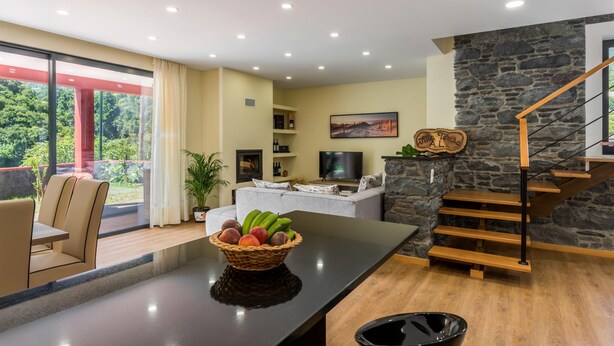 A modern living room with a large window, a dining area, a kitchen island with fruit, a TV, and a stone staircase. - Villa Teresa 1 by Atlantic Holiday (São Vicente)