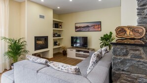 A living room with a grey sofa, a flat-screen TV, a stone fireplace, and a bookshelf with wine bottles. - Villa Teresa 1 by Atlantic Holiday (São Vicente)