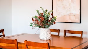 Una mesa de comedor con un florero blanco texturizado que contiene follaje verde y flores rojas.