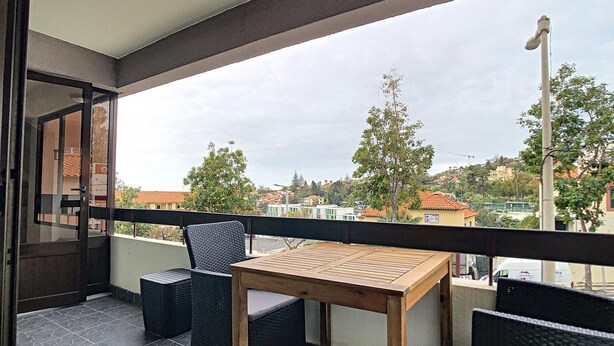 Un balcón con una mesa de madera y dos sillas de mimbre, con vistas a una zona residencial con casas y árboles.