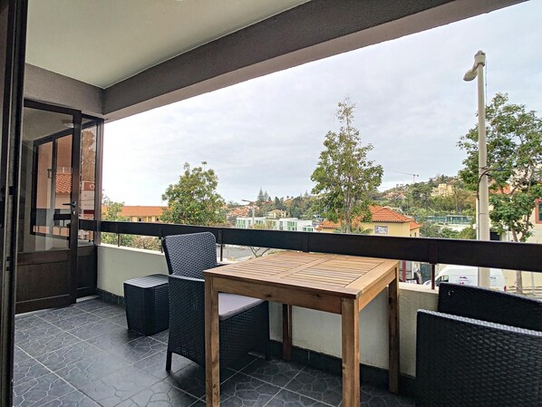Un balcón con una mesa de madera y dos sillas de mimbre, con vistas a una zona residencial con casas y árboles.