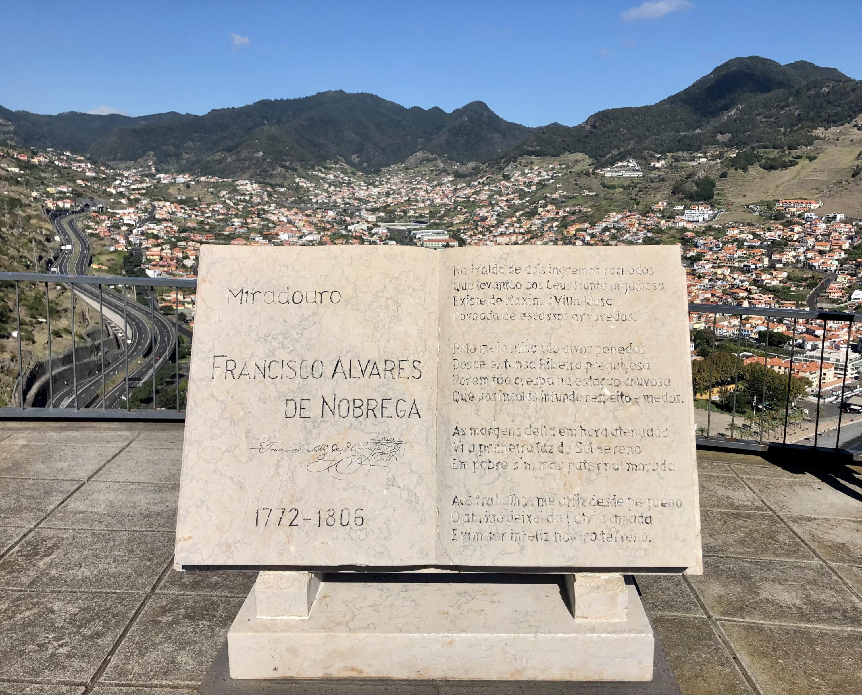 A memorial plaque with Francisco Alves de Nobrega's name and dates 1772-1806, set against a scenic backdrop of a winding road and a town nestled in hills.