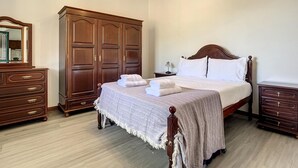 Un dormitorio con un amplio armario de madera, una cama con sábanas blancas, una mesita de noche y un aparador.