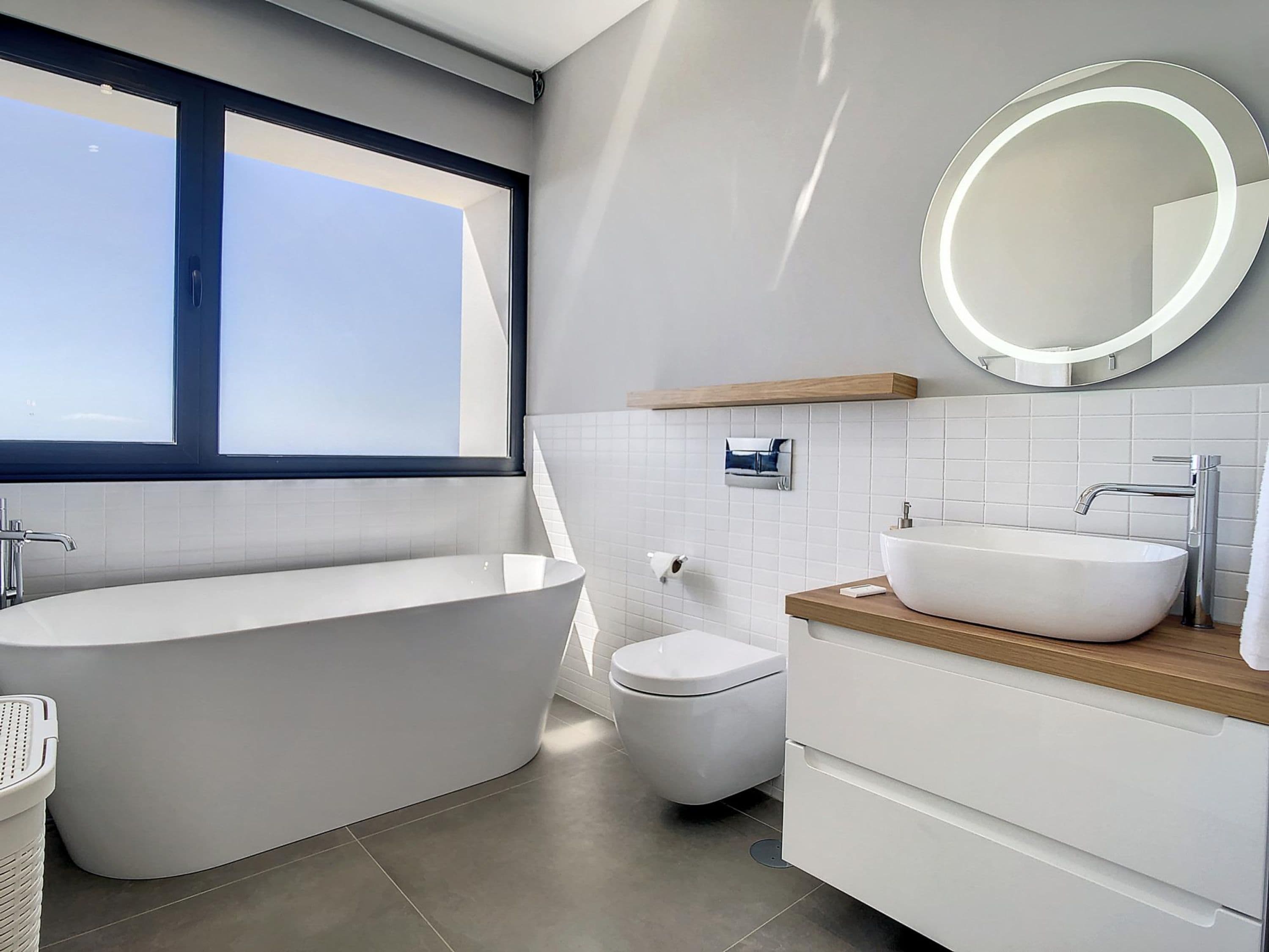 Een moderne badkamer met een vrijstaand bad, toilet, wastafel met spiegel en een raam voor natuurlijk licht.