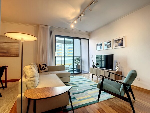 Una sala de estar moderna con un sofá, una mesa de centro, un televisor y un ventanal con vistas a edificios.