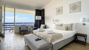 Habitación de hotel con una cama grande, un área de descanso con mesa y vistas al mar.
