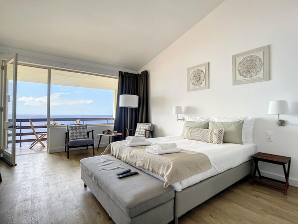 A hotel room with a large bed, a seating area with a table, and a view of the sea.