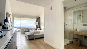 Una habitación de hotel moderna con una cama grande, baño independiente con ducha de vidrio, balcón con vistas y kitchenette.
