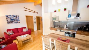 Een knus woongedeelte met een kitchenette, een rode bank, een houten eettafel en een wand met foto's.