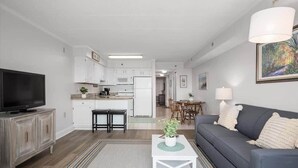 Una sala de estar moderna con televisor, sofá, mesa de centro y una cocina al fondo.