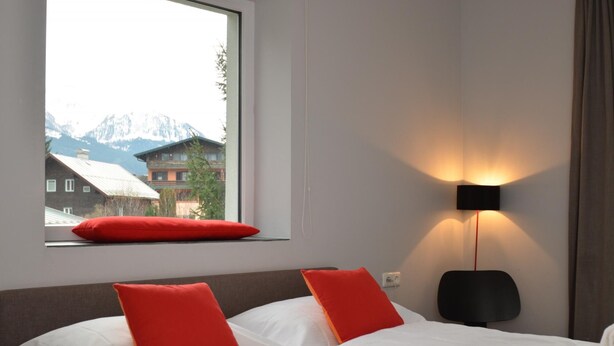 A hotel room with two beds, a window with a red pillow, a wall-mounted lamp, and a view of mountains. - 5 Seasons House Zell am See - TOP 4 Summer Card (Zell am See)