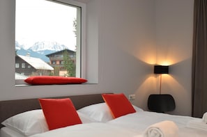 Habitación de hotel con dos camas, una ventana con almohada roja, una lámpara de pared y vistas a las montañas.