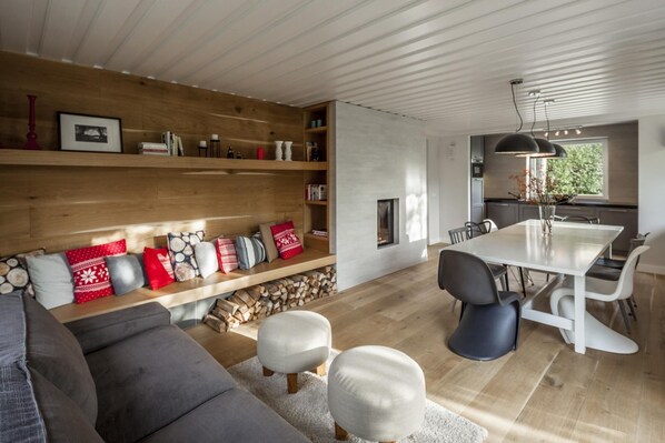 A modern living room with a grey sofa, white ottomans, a wooden bookshelf, and a dining area with a white table and chairs.