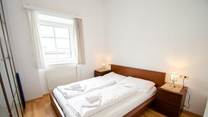 A single-bed room with a wooden headboard, white bedding, two bedside tables with lamps, a window with curtains, and a radiator. - Apartment Cityxpress TOP 7 - Zell am See (Zell am See)