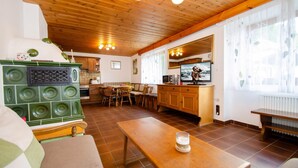 Ein gemütliches Wohnzimmer mit Holzbalkendecke, einem grünen Ofen, einem Essbereich und einer Küche mit Fernseher.