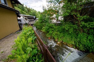 Un pequeño arroyo serpentea por una zona verde y exuberante, con un puente de madera y edificios al fondo.