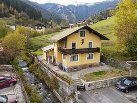 Una casa amarilla con balcón, una cascada pequeña y montañas al fondo.