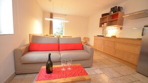 Una sala de estar con un sofá, una pequeña mesa de madera con una botella y vasos, una zona de cocina con armarios y un refrigerador, y una ventana que proporciona luz natural.