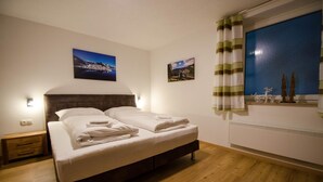 Una cama bien hecha con sábanas y toallas blancas, cabecera de madera y dos lámparas fijadas a la pared.