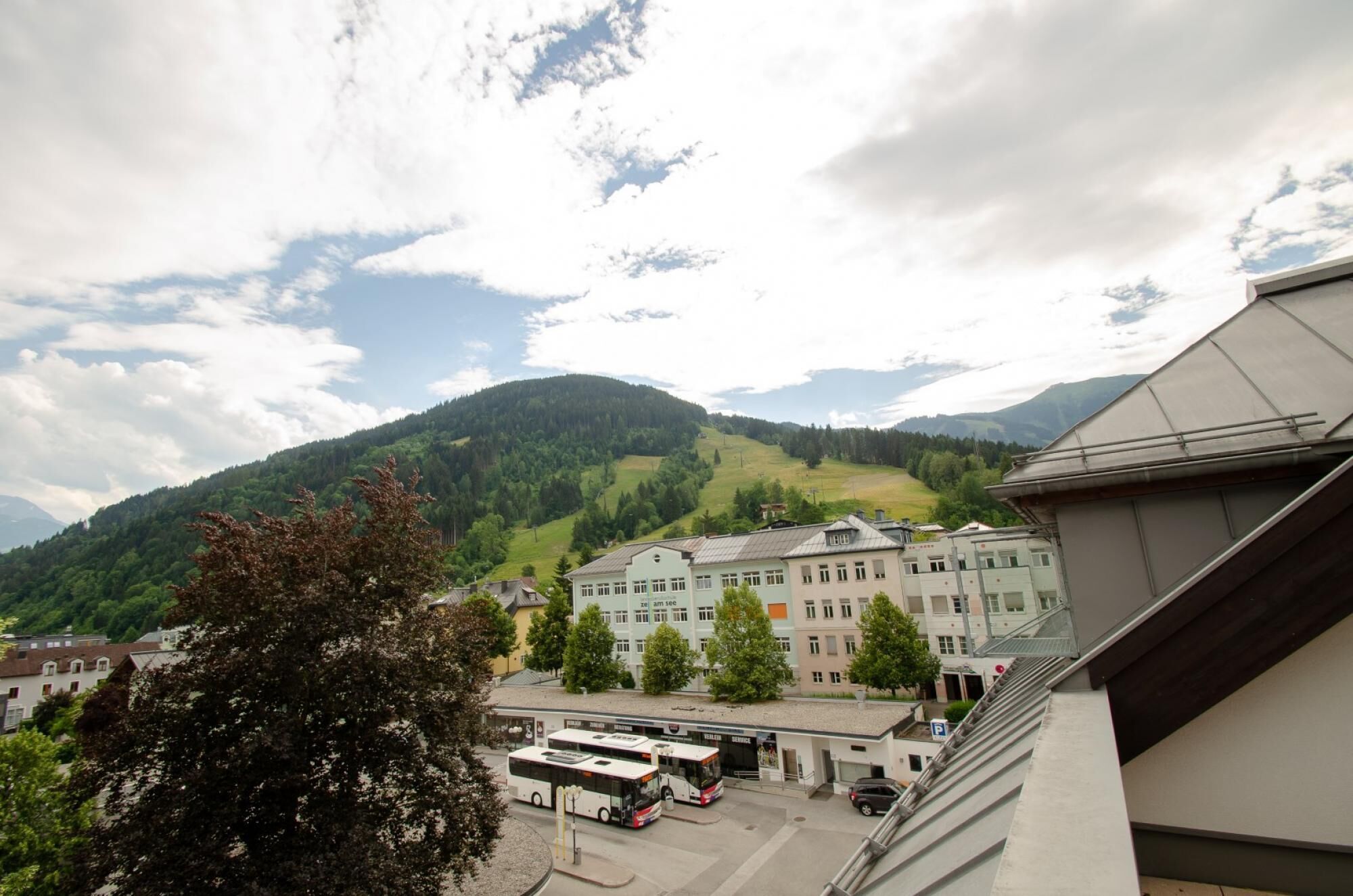 A bus station with a building in the background, surrounded by greenery and mountains.
