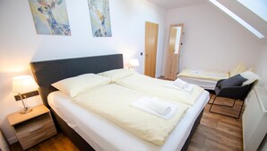 Une chambre d’hôtel avec un grand lit, un plus petit lit, un bureau et une chaise. Il y a des tableaux au mur et une porte sur la droite.
