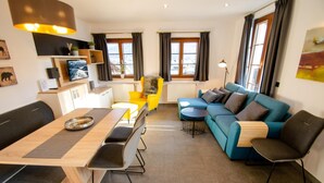 Un salon moderne comprenant un coin repas, un canapé et un fauteuil. On y trouve des fenêtres avec des rideaux, une lampe et un tableau au mur.